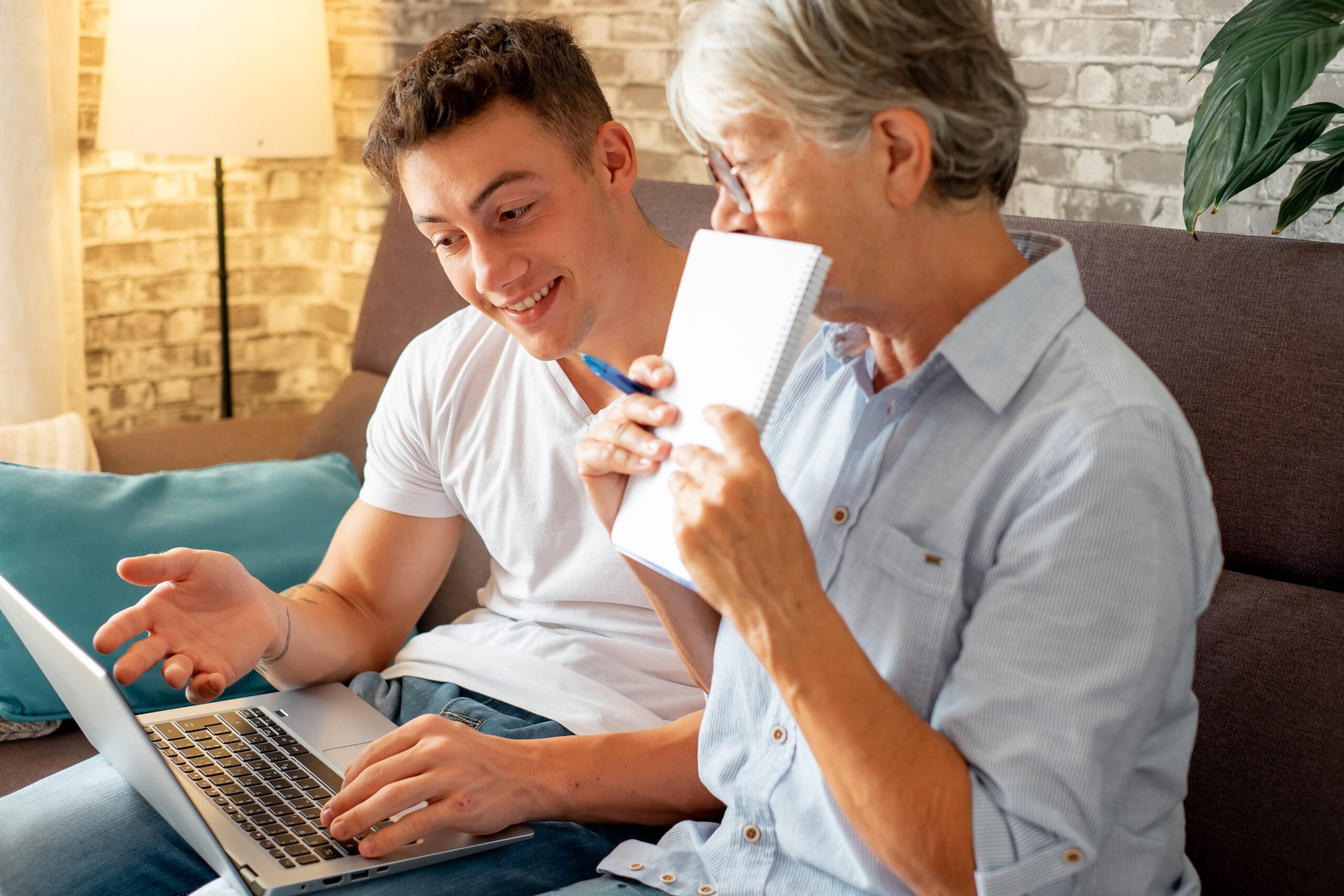 Beautiful young boy sitting on sofa at home helping senior grandmother surf with laptop on the web while she takes notes. Younger generations care for older relatives teaching how to use computer